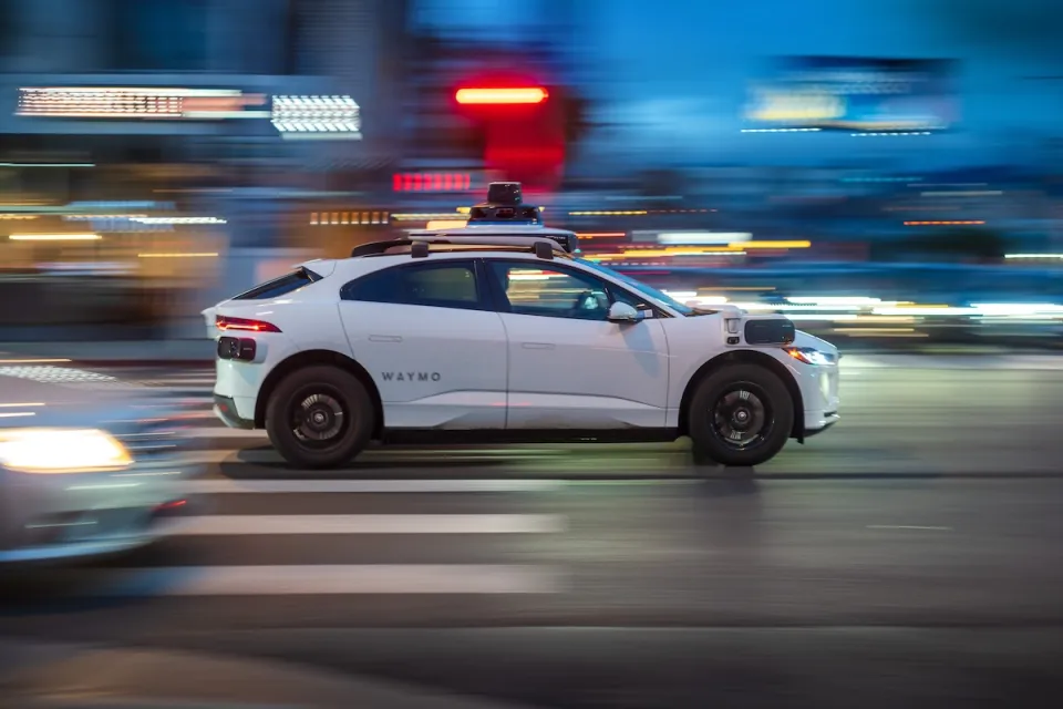 White Waymo autonomous car driving fast down city street with blurred background at night.